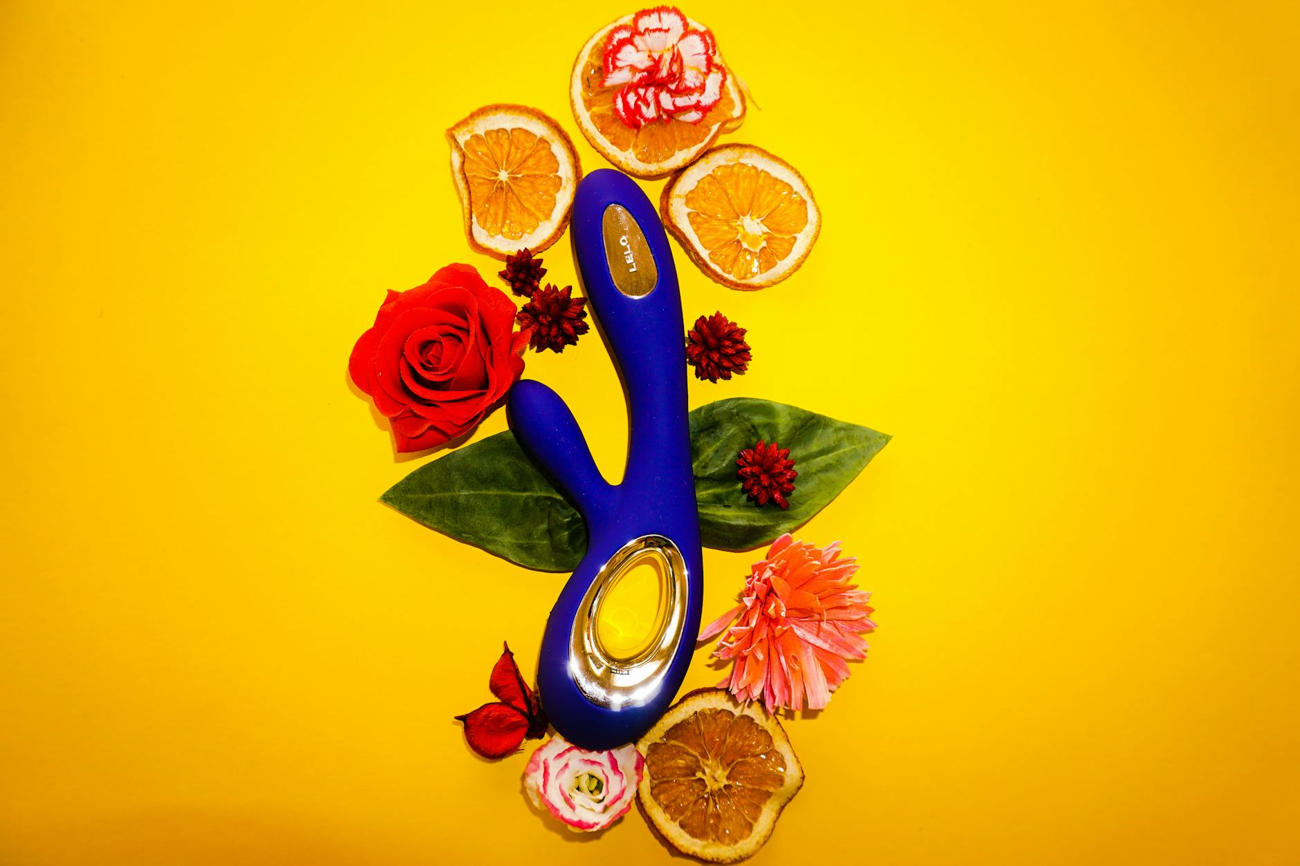 A blue clitoral vibrator surrounded by flowers and citrus on a bright background